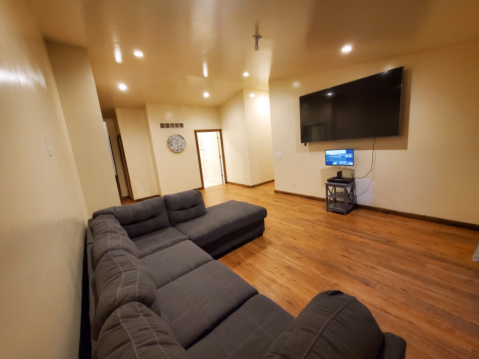 A spacious living room with wooden flooring, beige walls, and recessed ceiling lights. There is a large gray sectional sofa on the left side and a large flat-screen TV mounted on the wall opposite the sofa. Below the TV is a small stand with a smaller monitor and some electronic devices. A doorway and a wall clock are visible in the background.