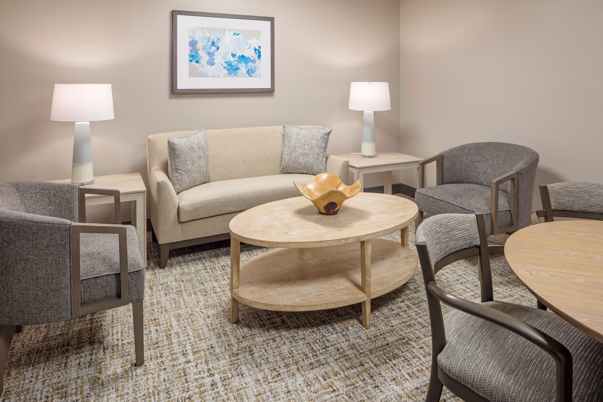 Small sitting room with a beige sofa, gray armchairs, a round wooden coffee table, table lamps, and framed artwork on the wall.