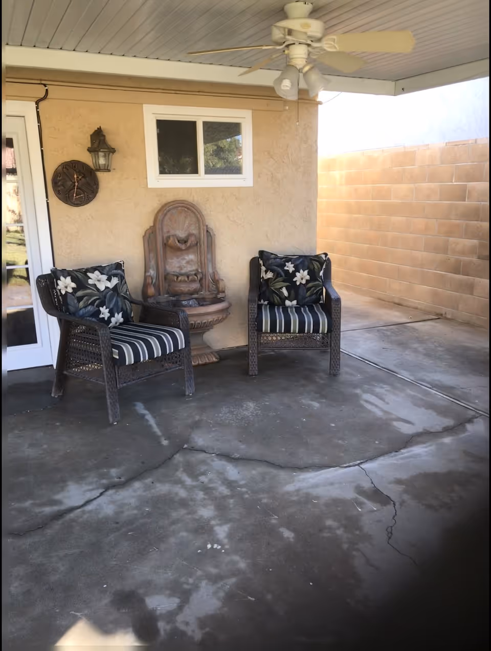 Covered patio with two wicker chairs with floral cushions, a wall-mounted fountain, and a ceiling fan.