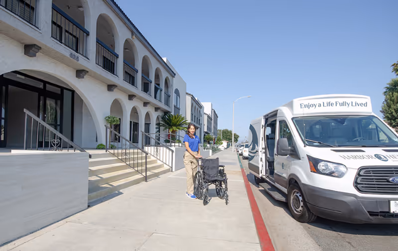 Harbor Heights exterior with arched walkway, sidewalk, and a staffer beside a wheelchair.