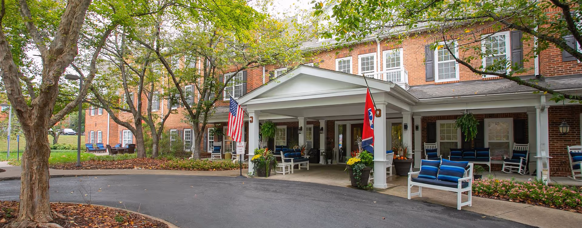 Front entrance of a brick senior living building with a covered porte-cochere, flags, benches and trees.