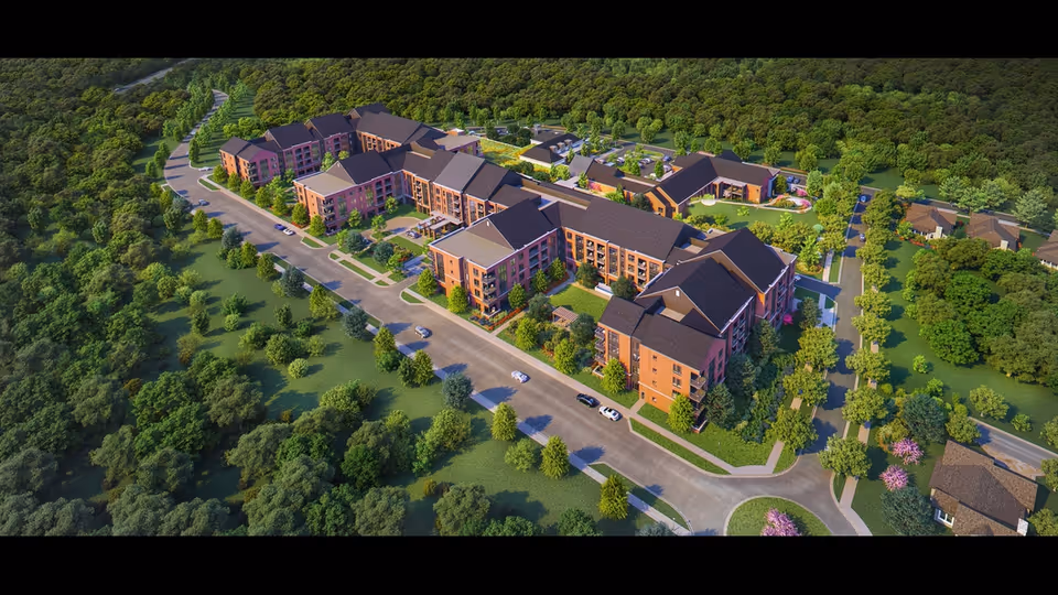 Aerial view of The Village at Providence Point senior living facility showing multiple connected buildings with pitched roofs surrounded by trees and greenery, with roads and parked cars visible around the complex.