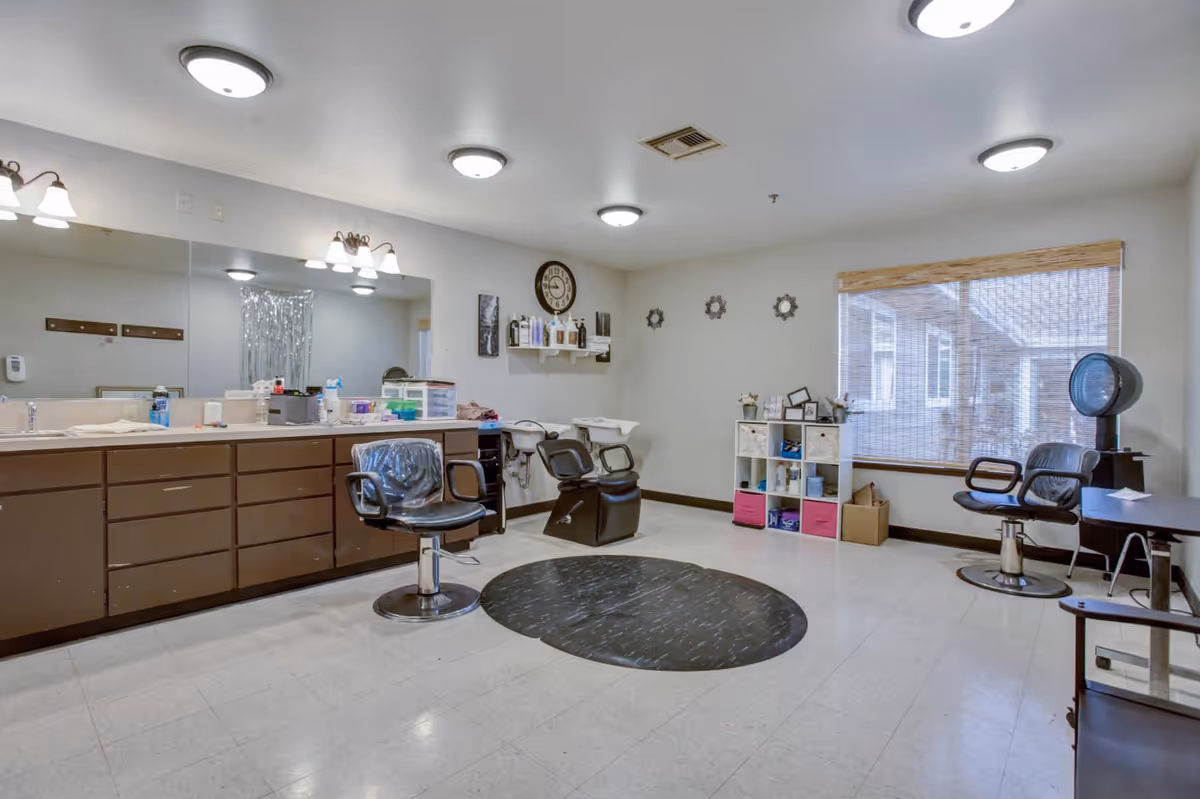 A spacious salon room with styling chairs, wash stations, a large mirror, and storage cabinets in a senior living facility.