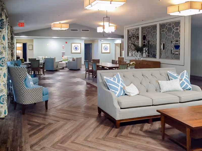 A spacious and well-lit common area in a senior living facility featuring a beige tufted sofa with patterned blue and white pillows, a wooden coffee table, several armchairs, and dining tables with chairs. The room has wood-patterned flooring, decorative wall panels, framed artwork, and modern ceiling lights. An exit sign is visible on the far wall.