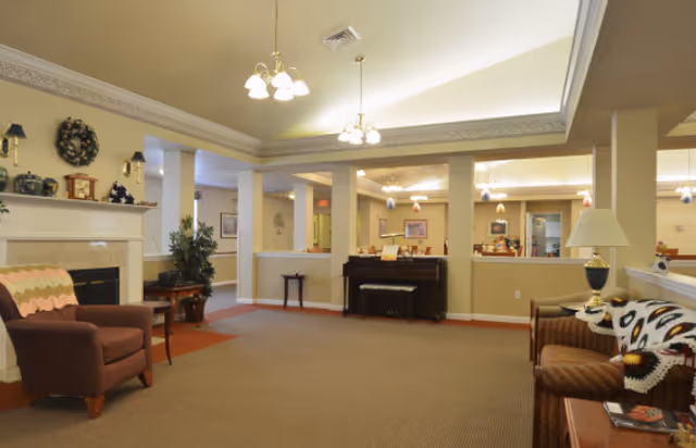 A spacious living room area in a senior living facility with beige walls and carpet. The room features a brown armchair with a crocheted blanket, a fireplace with decorative items on the mantle, a piano against the far wall, and a couch with a patterned throw. There are ceiling lights and a table lamp providing warm lighting, and the room has an open layout with visible adjoining rooms.