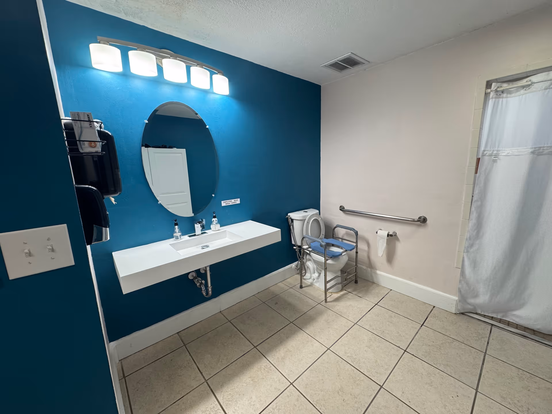 Accessible bathroom with a wall-mounted sink and round mirror, a raised-seat toilet with a support frame and grab bar, and a shower with a curtain.