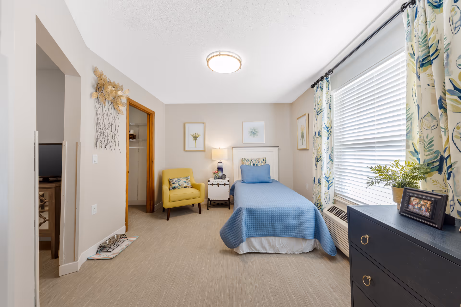 Well-lit bedroom with a twin bed covered in a blue quilt, a yellow armchair, dresser, and a large window with floral curtains.