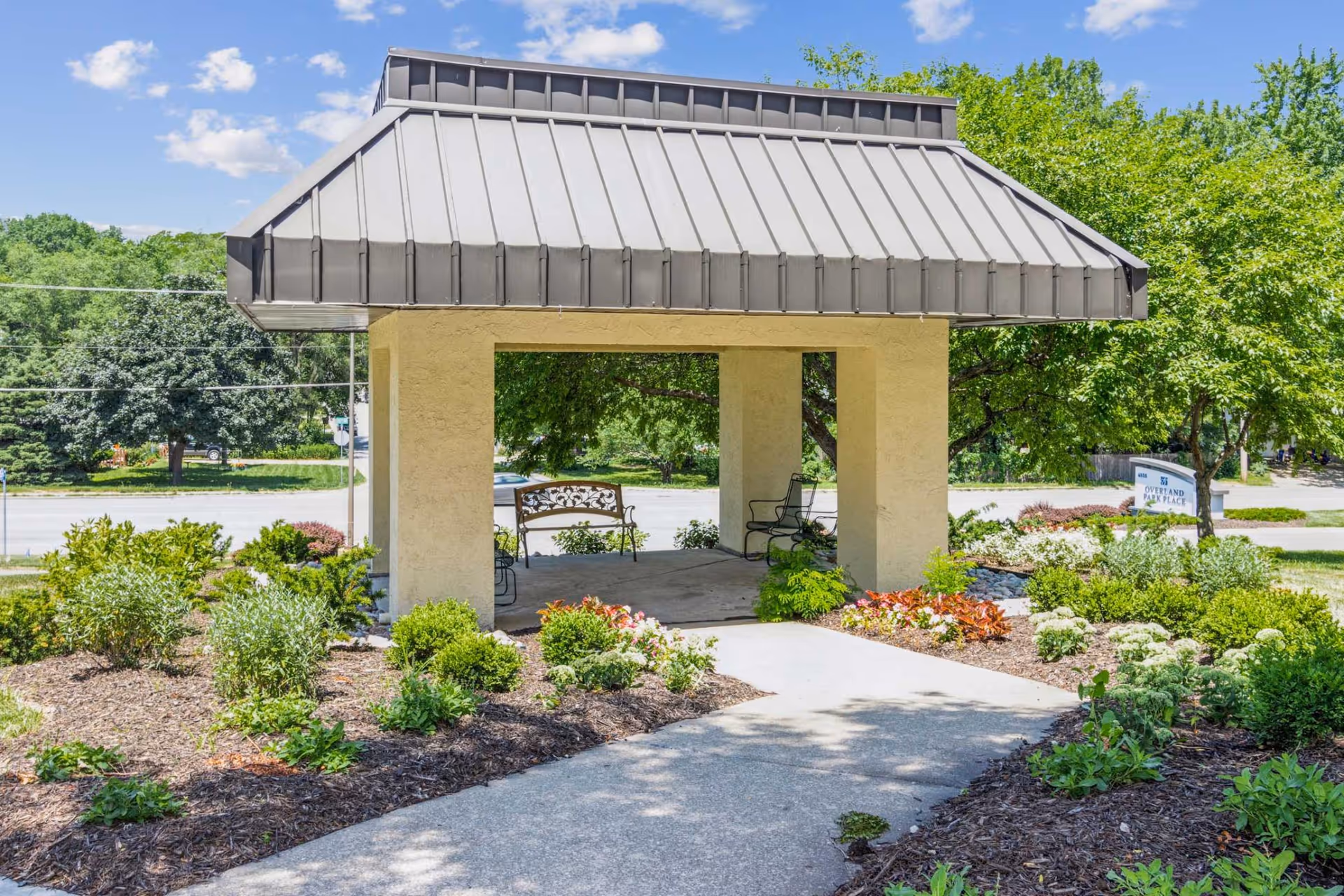 Covered entrance pavilion with benches, a walkway, and landscaped garden beds in front of a senior living facility.