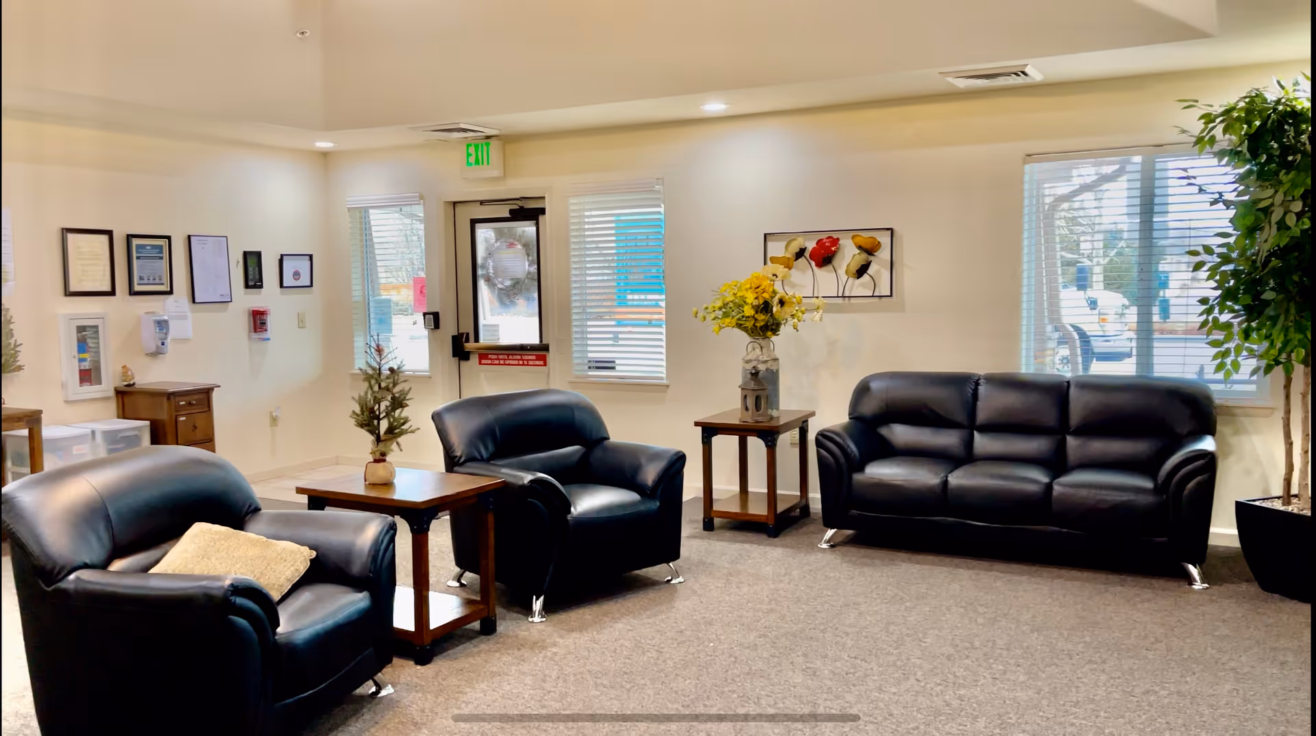 A cozy living room area in Aspen House featuring two black leather armchairs and a black leather sofa arranged around two wooden side tables. One table has a small plant, and the other has a vase with yellow flowers. The room has beige walls with framed certificates and artwork, windows with blinds, a door with an exit sign above it, and a large potted plant in the corner.