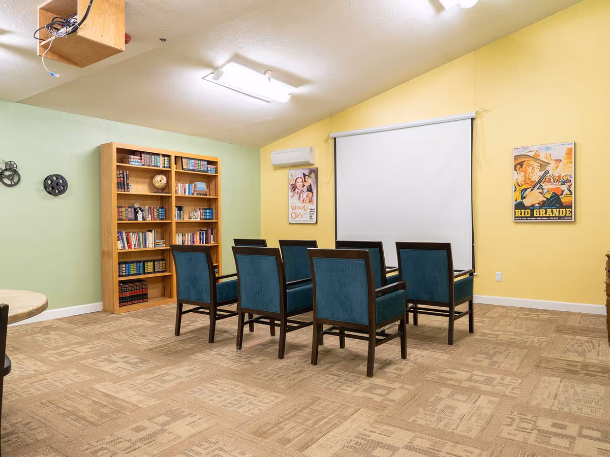 A small meeting or media room with six blue upholstered chairs arranged in two rows facing a white projection screen on a yellow wall. There is a wooden bookshelf filled with books against a light green wall, and two vintage movie posters, one for 'Wizard of Oz' and another for 'Rio Grande', hang on the yellow wall. The room has beige patterned carpet and a ceiling-mounted projector.