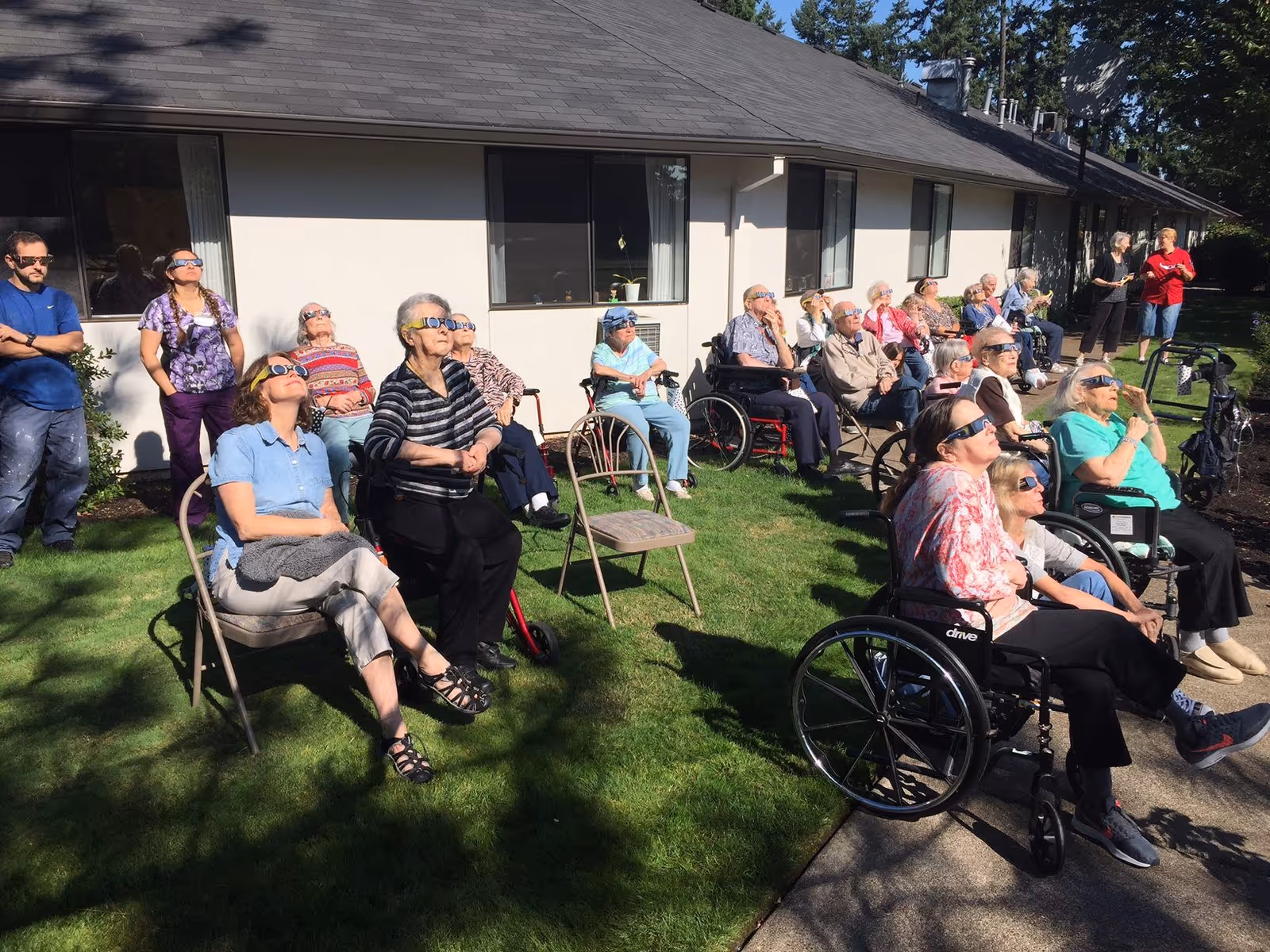 A group of elderly people, some seated in wheelchairs and others in chairs, gathered outside on a lawn next to a building. They are all wearing protective eclipse glasses and looking up at the sky, likely watching a solar eclipse. Two staff members stand nearby observing the group.