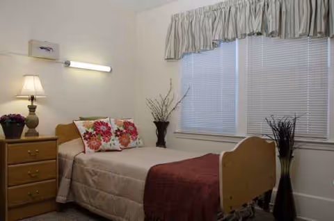 A bedroom in a senior living facility with a bed, dresser, and window blinds.