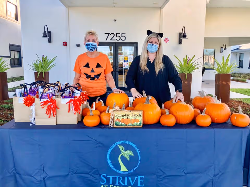 Two women wearing face masks stand behind a table covered with a blue tablecloth that has the Strive at Fern Park logo. The table is filled with various sizes of pumpkins and gift bags decorated with red and white ribbons. One woman is wearing an orange shirt with a jack-o'-lantern face, and the other is dressed in black with cat ears. They are standing outside in front of a building entrance with the number 7255 above the door.