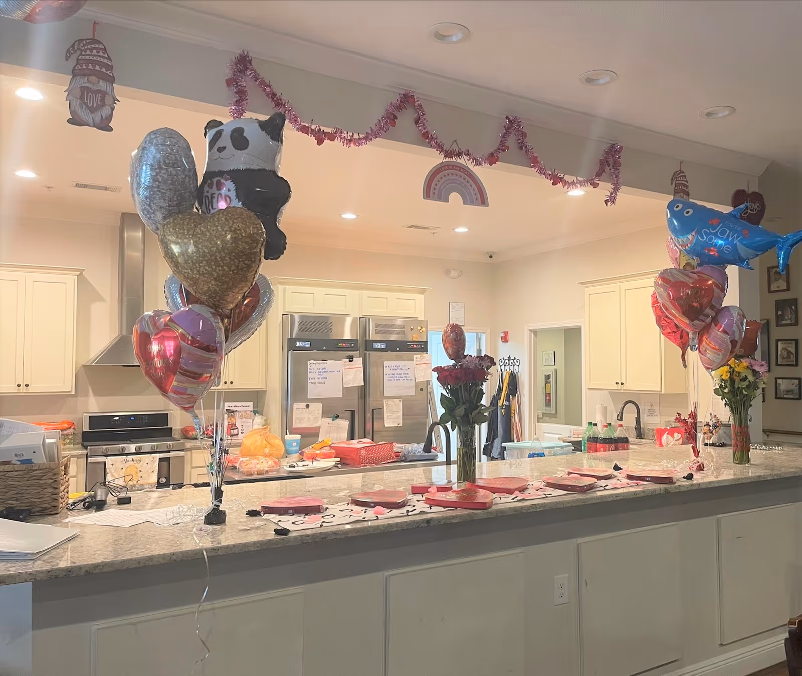 A kitchen area decorated with Valentine-themed balloons, including heart shapes, a panda, and a shark. The countertop has several pink and red plates arranged with napkins and utensils. There are flowers in vases on the counter and pink garland hanging from the ceiling. The kitchen has white cabinets, stainless steel appliances, and a refrigerator with notes attached.