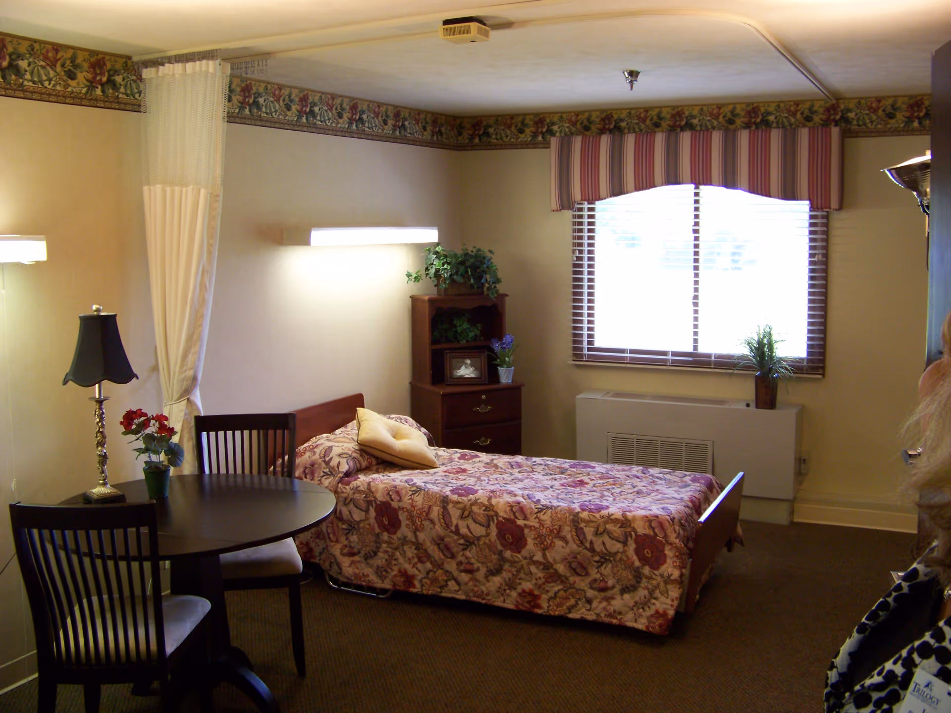A small, cozy room with a single bed covered in a floral bedspread, a wooden nightstand with plants and a framed photo, a round table with two chairs, a lamp, and a window with striped valance and blinds. The room has beige walls with floral wallpaper border near the ceiling and a privacy curtain hanging from the ceiling.