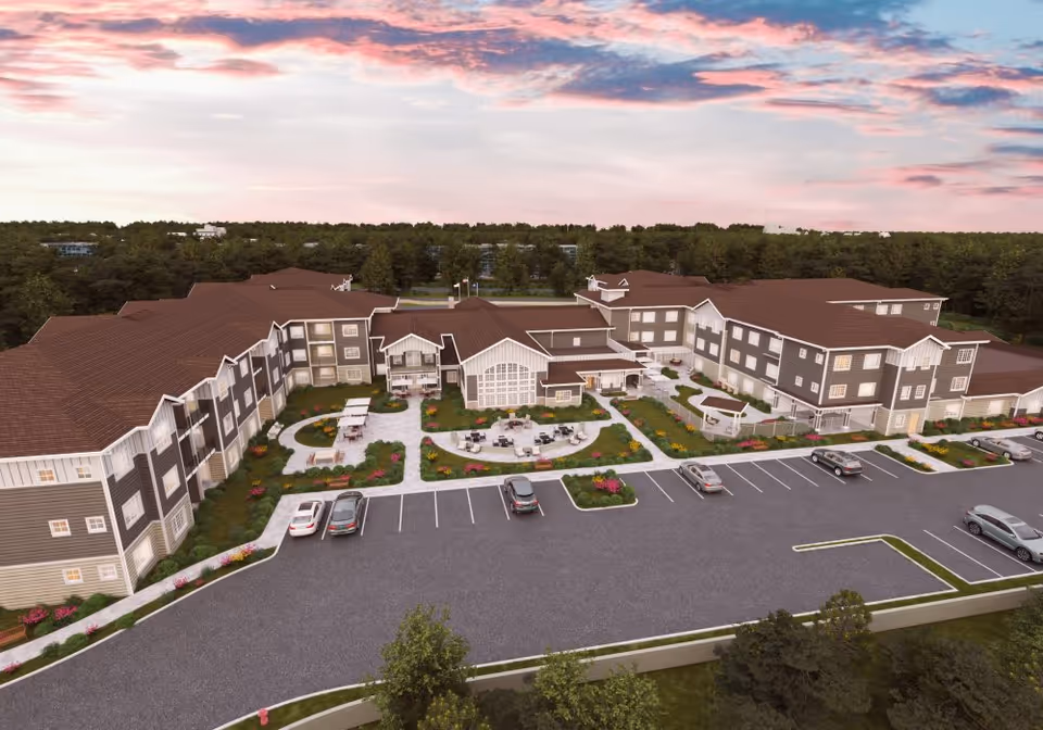 Aerial view of a large senior living facility building with brown roofs and gray siding, surrounded by landscaped gardens and outdoor seating areas. Several cars are parked in the parking lot in front of the building, and trees and greenery extend into the background under a colorful sky at sunset.