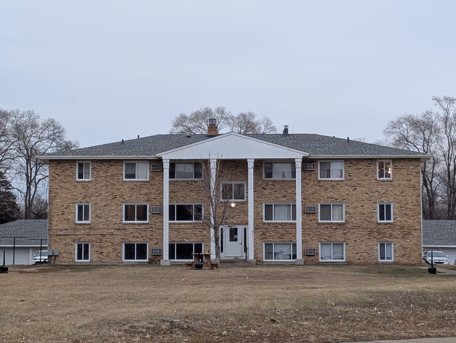 Brick three-story apartment-style building with white columns and a central entrance set on a grassy lawn.