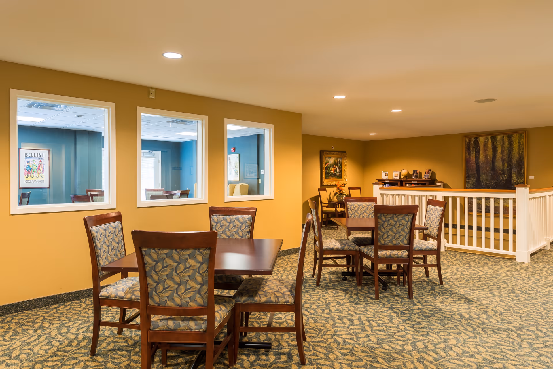 A cozy interior common area with multiple wooden tables and chairs featuring patterned upholstery. The walls are painted a warm yellow, and there are three windows looking into an adjacent room with blue walls. The carpet has a leafy pattern, and there are framed paintings on the walls. A white railing separates part of the space.