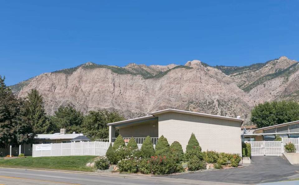 Single-story light-brick building with manicured shrubs and a white fence in front of a large mountain under a clear blue sky.