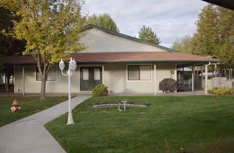 Single-story building with a sloped roof, surrounded by green grass and trees. A concrete walkway leads to a green double door entrance. There is a white lamp post near the walkway and a fire hydrant on the grass. The building has a covered porch area with outdoor furniture and a barbecue grill covered with a black cover.