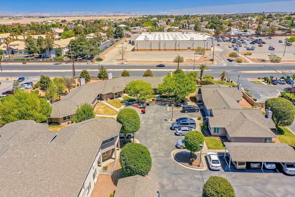 Aerial view of Solstice Senior Living at Rio Norte facility showing multiple single-story buildings arranged around a parking lot with several cars parked. The area is landscaped with green trees and bushes, and a road with more buildings and parking lots is visible in the background under a clear blue sky.
