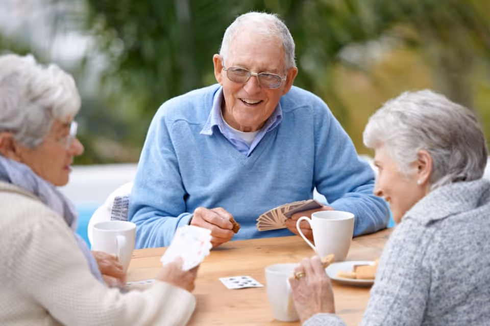 Three elderly people sitting around a wooden table outdoors, playing cards and enjoying snacks and drinks, with greenery in the background.