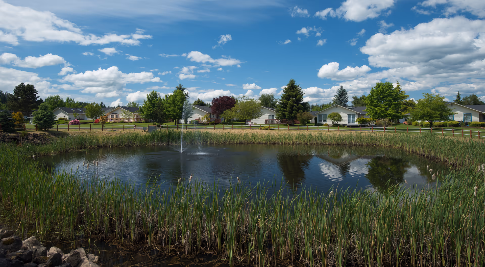 A small pond with a central fountain in front of single-story homes, trees, and a blue sky with scattered clouds.