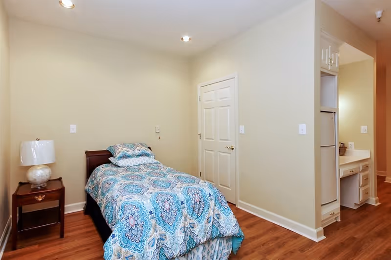 A small bedroom with a single bed covered in a blue and white patterned bedspread. Next to the bed is a wooden nightstand with a white lamp. The room has light-colored walls and wooden flooring. There is a closed white door on the far wall and a small kitchenette area with a refrigerator and countertop visible to the right.