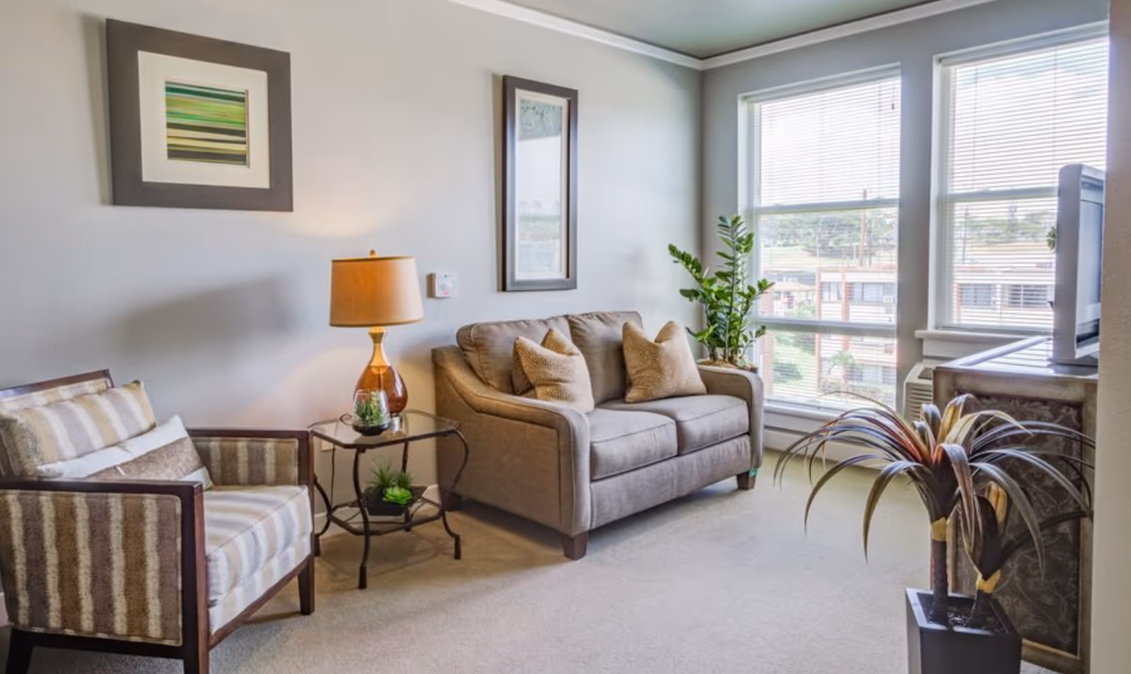 A bright living room with a loveseat, armchair, side table with a lamp, potted plants, and large windows.