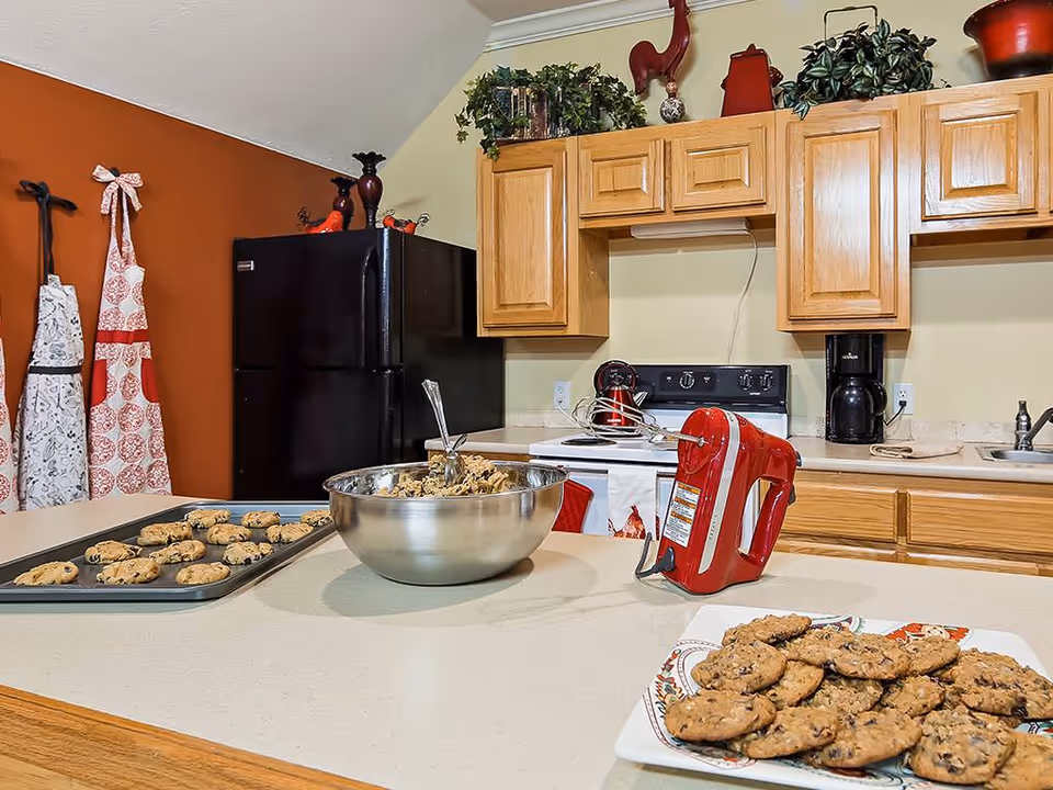 A kitchen with wooden cabinets, a black refrigerator, a stove, and a coffee maker. On the counter, there is a red hand mixer, a large metal mixing bowl with cookie dough and a spoon, a baking tray with unbaked cookies, and a plate with baked cookies. Two aprons hang on the wall, and decorative items are placed on top of the cabinets.