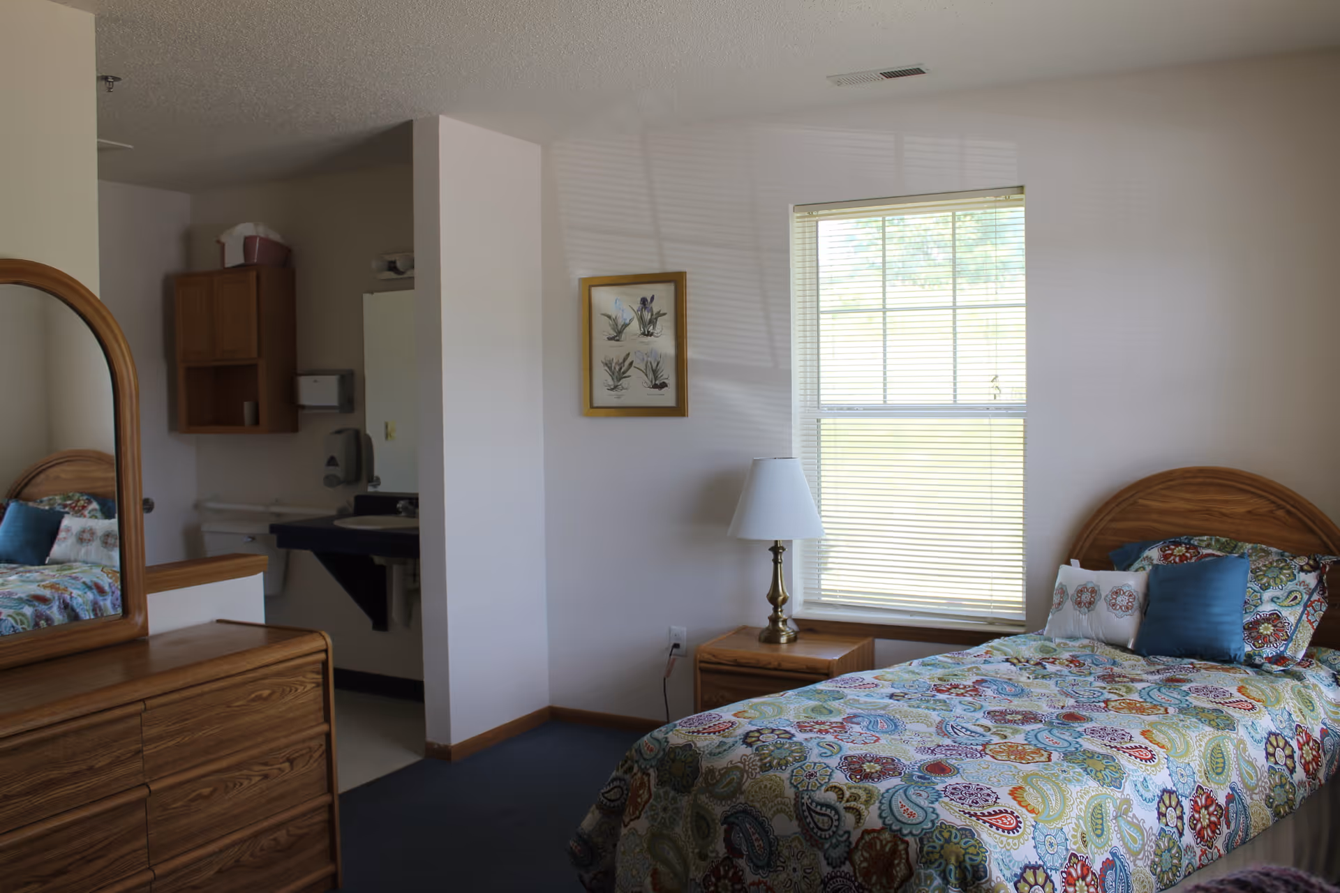 Single-bed bedroom with wooden furniture, a patterned bedspread, a nightstand and lamp, dresser with mirror, and a small sink area visible through an open partition.