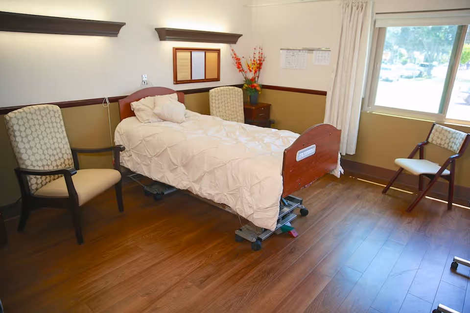 Private senior-care bedroom with a raised hospital bed, two chairs, a nightstand with flowers, and a large window.