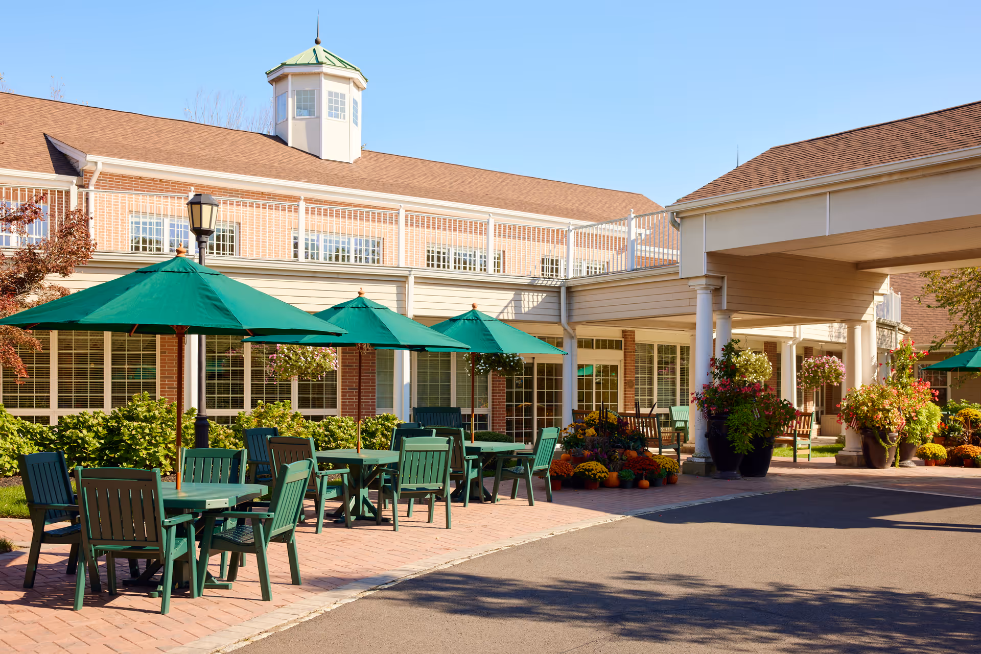 Outdoor seating area at Brandywine Colts Neck by Monarch with green tables and chairs under green umbrellas, surrounded by potted plants and flowers, adjacent to a brick building with large windows and a covered entrance.