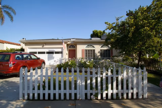 Single-story residential house with a white picket fence in front, a red car parked in the driveway, an American flag near the front door, and trees and bushes surrounding the property under a clear blue sky.