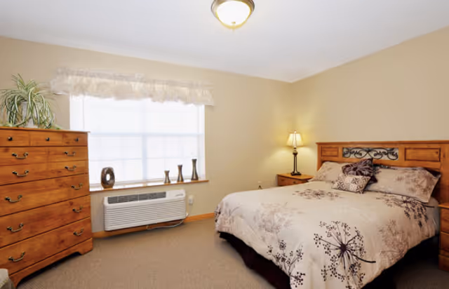 Bright bedroom with a patterned bed, wooden dresser, nightstand with lamp, and a window air conditioner.