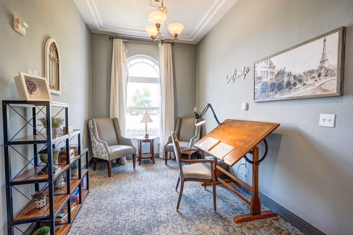 A cozy interior room with a large arched window covered by white curtains, two upholstered armchairs with a small wooden side table and lamp between them, a wooden drafting table with a chair and a desk lamp, a metal and wood shelving unit with decorative items, and wall art including a 'welcome' sign and a framed picture of a cityscape with the Eiffel Tower.