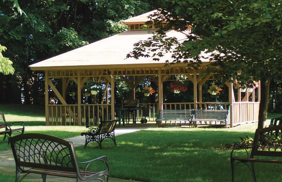A wooden gazebo in a green outdoor area with hanging flower pots and several metal benches placed on the grass around it, surrounded by trees.