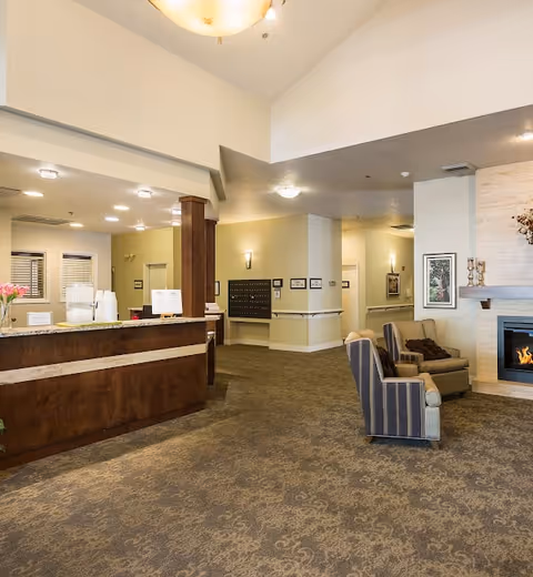 Interior view of a senior living facility lobby with a wooden reception desk on the left, a seating area with armchairs and a fireplace on the right, beige walls with framed artwork, and a carpeted floor.