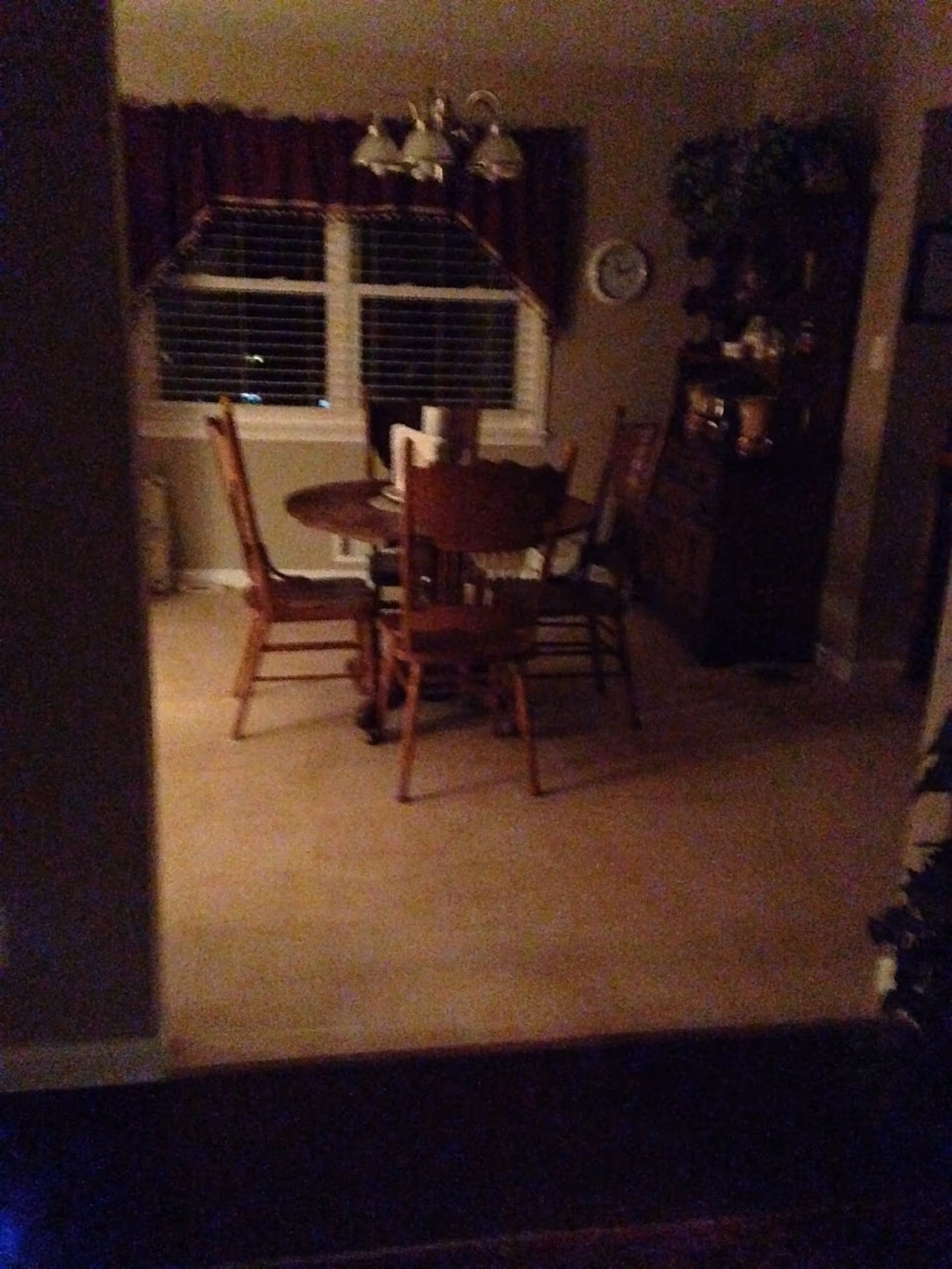 Dimly lit dining area with a round wooden table surrounded by four wooden chairs. A chandelier with multiple lights hangs above the table. Behind the table is a window with closed blinds and a valance. To the right, there is a tall wooden cabinet with decorative items and a clock on the wall.