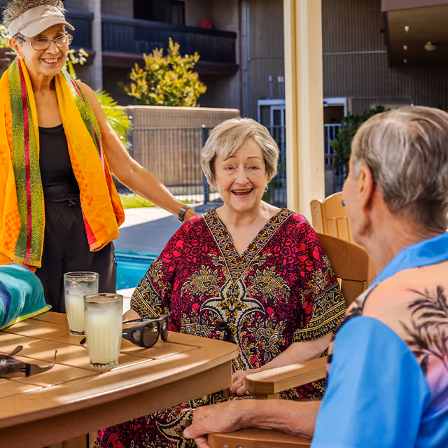 Three elderly individuals enjoying a sunny day by a pool at a retirement community. One woman wearing a colorful dress is seated and smiling, another woman with a visor and a bright towel around her neck stands nearby, and a man in a tropical shirt sits across the table. There are two glasses of lemonade and sunglasses on the table.