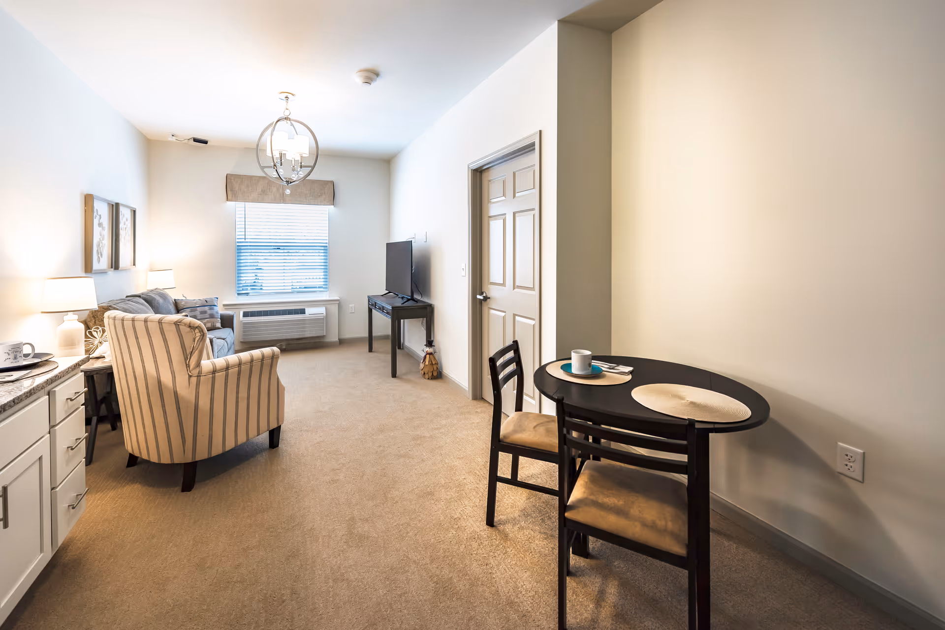 A cozy living area in a senior living facility featuring a striped armchair, a gray sofa, a TV on a stand, and a small round dining table with two chairs. The room has beige carpeting, white walls, a window with blinds, and soft lighting from table lamps and a ceiling light fixture.