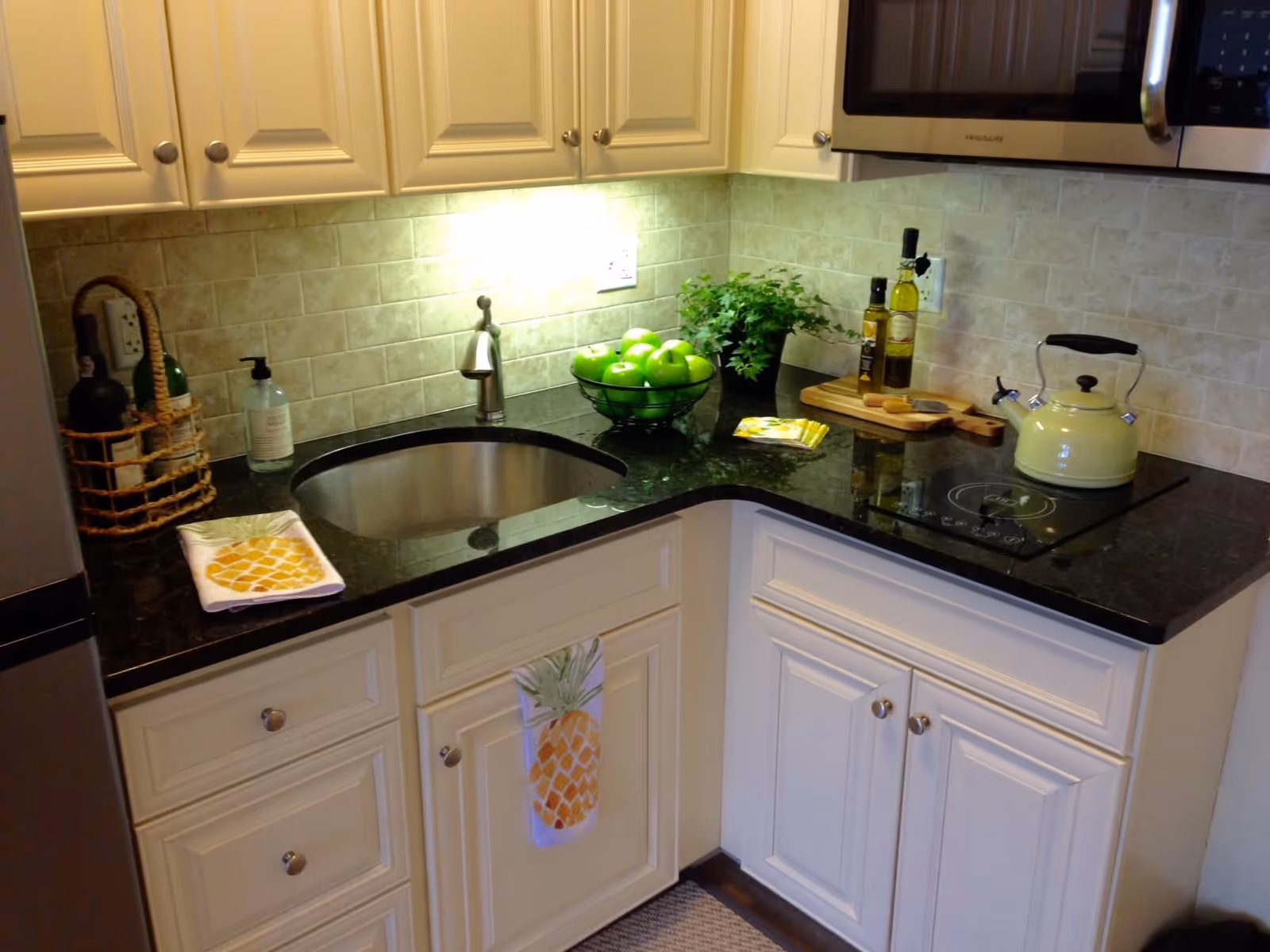 A small kitchen corner with white cabinets and black granite countertops. There is a stainless steel sink with a modern faucet, a bowl of green apples, a potted plant, bottles of olive oil, a yellow kettle on an electric stovetop, and a microwave above. A pineapple-themed towel hangs on one cabinet door.
