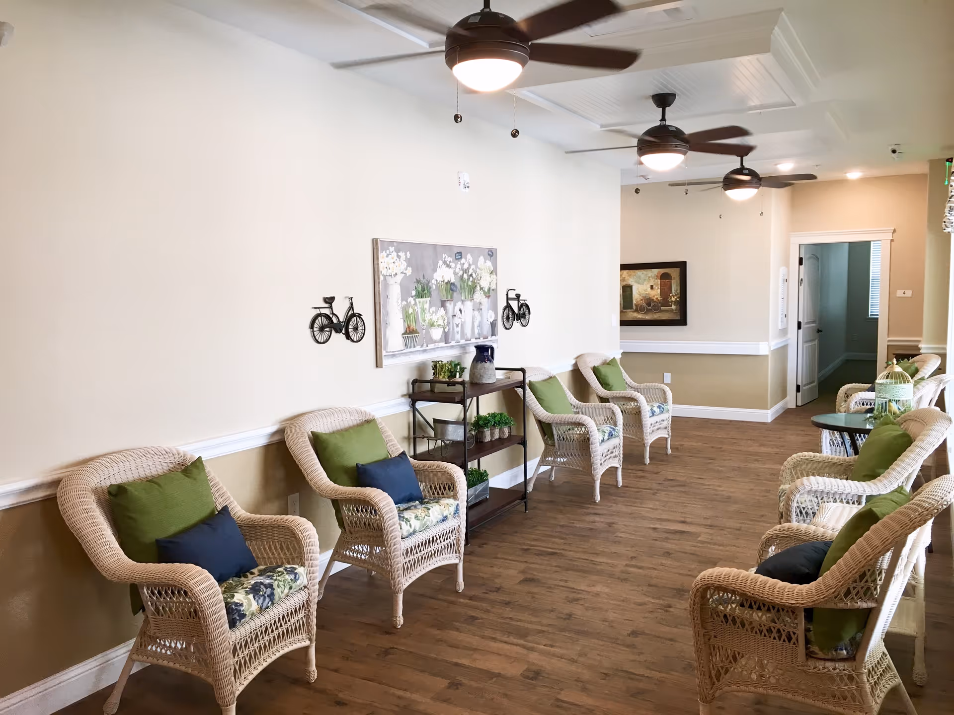 Bright communal seating area with wicker chairs, green and blue cushions, wood floor, ceiling fans, and wall art.