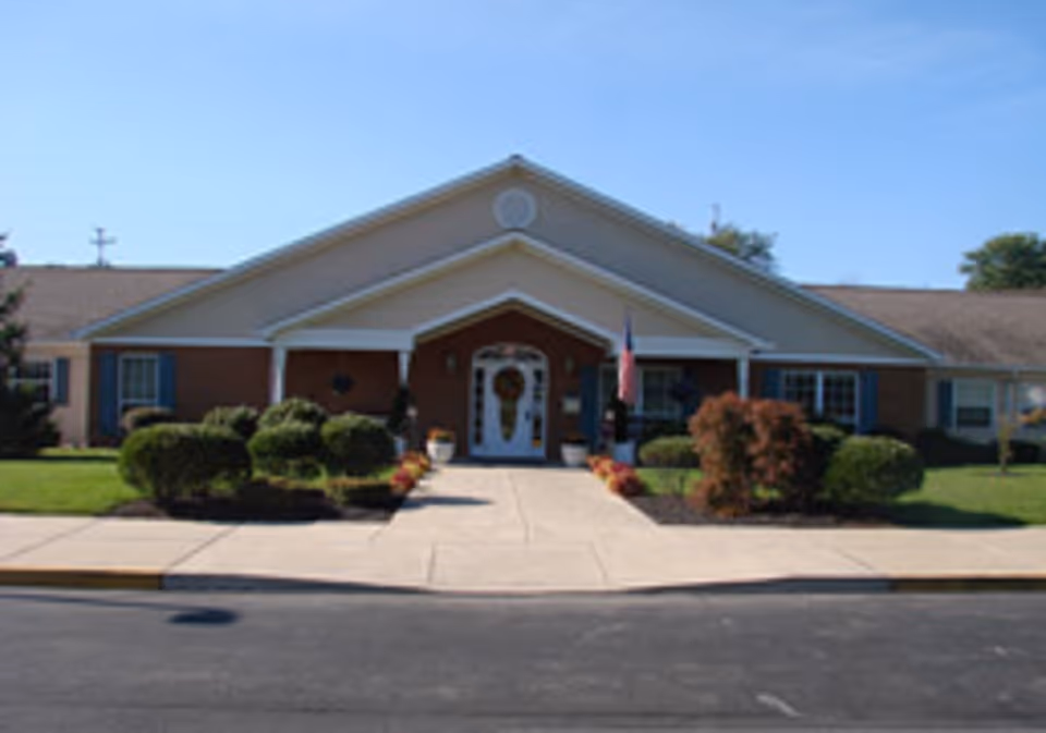 Single-story brick building with a peaked central entrance, a sidewalk leading to the door, landscaped shrubs, and an American flag.