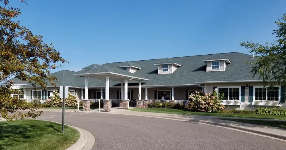 Front exterior of a single-story senior living building with a covered entrance, porch, and landscaped grounds under a clear sky.