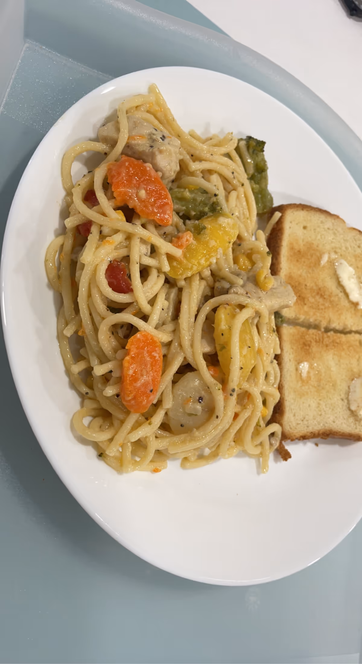 A white plate with spaghetti pasta mixed with vegetables including carrots, broccoli, and yellow bell peppers, served with two slices of buttered toast on the side.