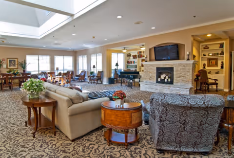 Spacious living room area with patterned carpet, beige and patterned armchairs, wooden side tables with plants and flowers, a stone fireplace with candles, a mounted flat-screen TV above the fireplace, large windows letting in natural light, and built-in shelves with decorative items.