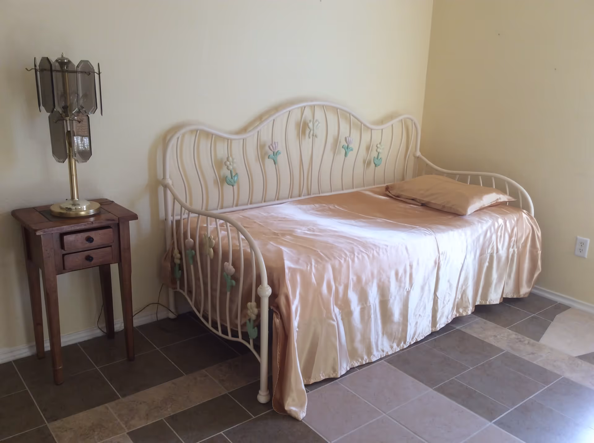 A light-colored metal daybed with floral accents covered in a silky peach bedspread beside a wooden nightstand and lamp in a tiled room.