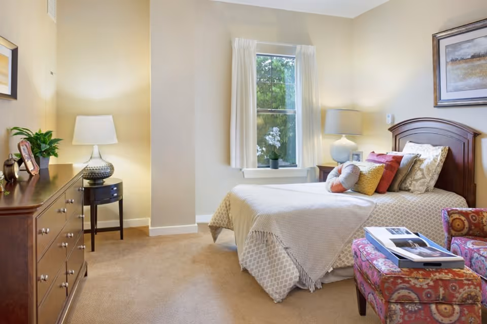 A cozy bedroom with a single bed adorned with multiple pillows and a beige patterned blanket. There is a window with white curtains letting in natural light. To the left, there is a wooden dresser with a lamp, a plant, and a framed photo. A small round side table with a lamp is next to the dresser. In front of the bed, there is a colorful patterned ottoman with a tray holding an open book. The walls are painted a light beige color and there is a framed artwork hanging above the bed.