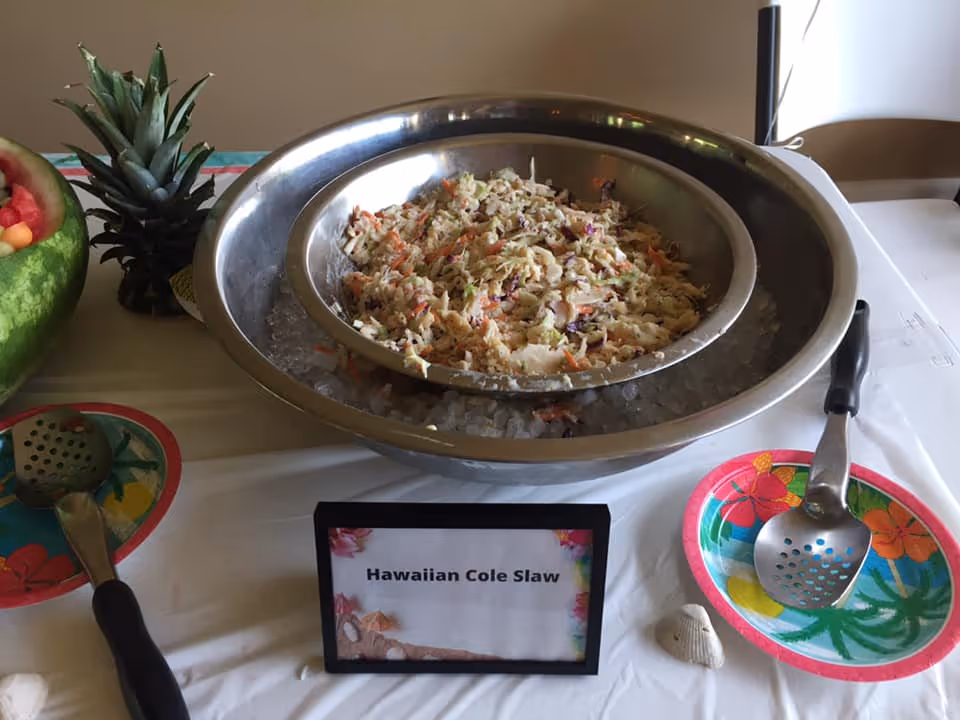 A serving station with a large metal bowl filled with Hawaiian cole slaw placed on ice. There are two serving spoons on colorful tropical-themed plates on either side of the bowl. A small sign in front of the bowl reads 'Hawaiian Cole Slaw'. A pineapple and a watermelon bowl filled with fruit are partially visible on the left side of the image.